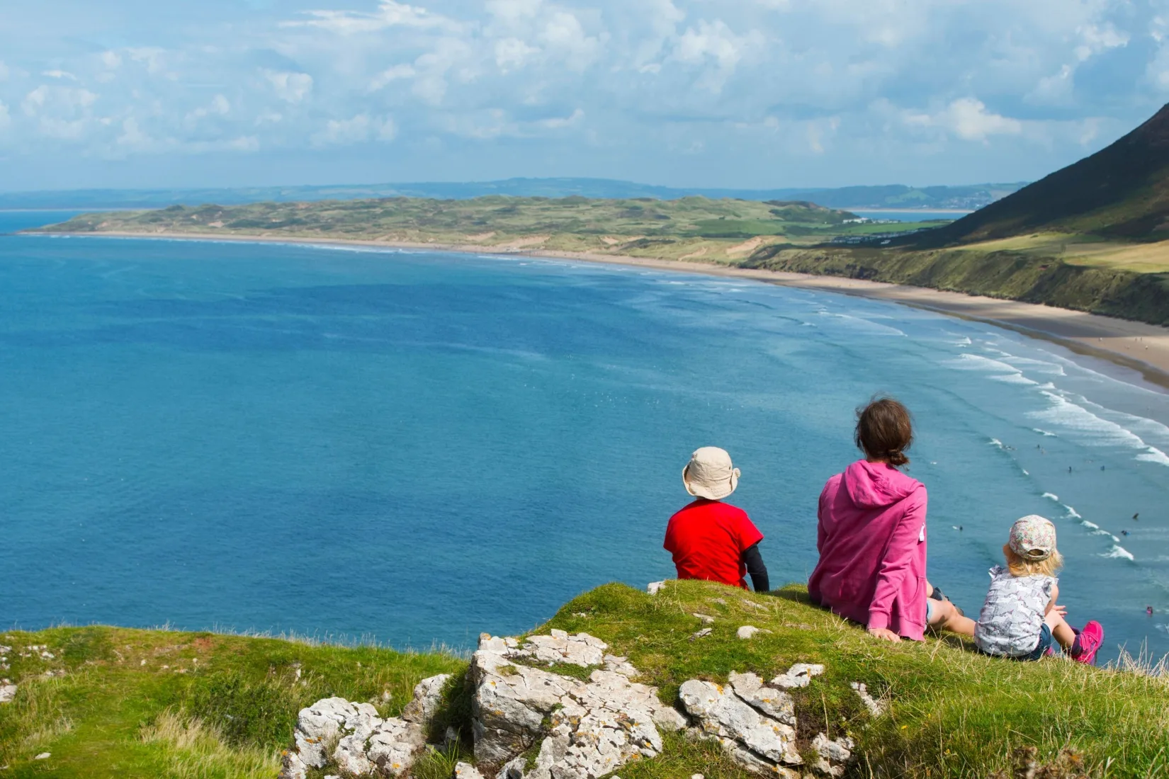 A family sitting on a clifftop overlooking a beach.